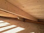 Das Foto zeigt einen Dachboden mit neuer Holzkonstruktion. Helle Balken stützen die Holzverkleidung des Dachs. Unten verläuft ein graues Rohr entlang der Wand. Sonnenlicht fällt durch eine Öffnung auf den staubigen Boden.